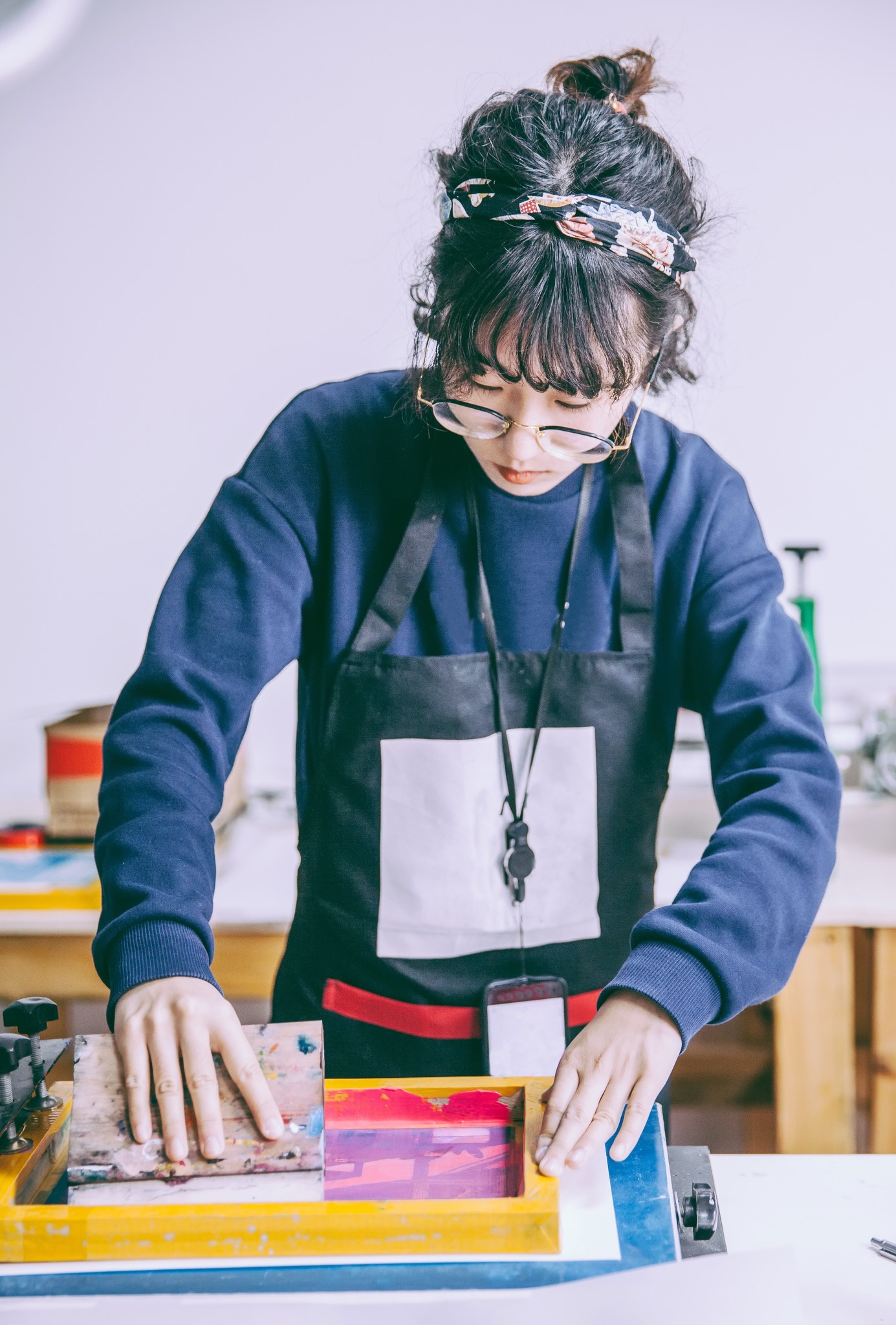 A woman wearing glasses and an apron pulls ink across a screen while working at a screen printing press.